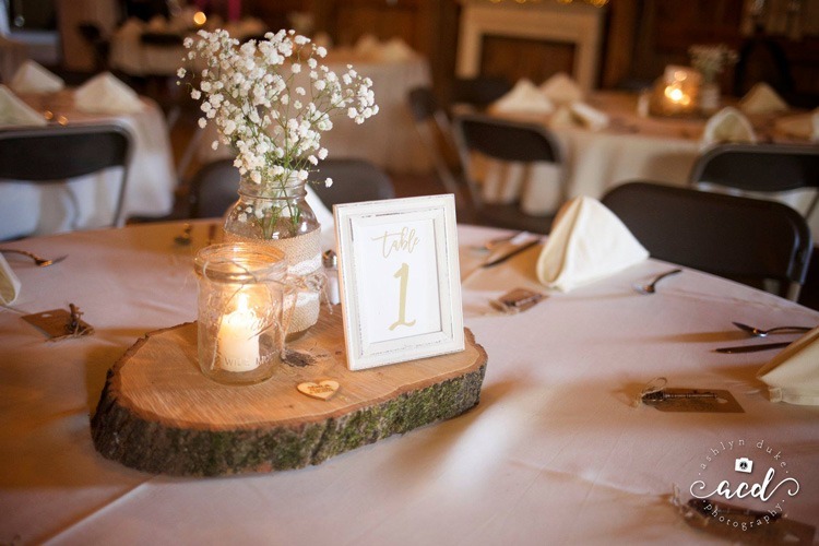 mason jar candle, mason jar vase with white florals, and table number grouped on piece of wood as centerpiece for barn wedding